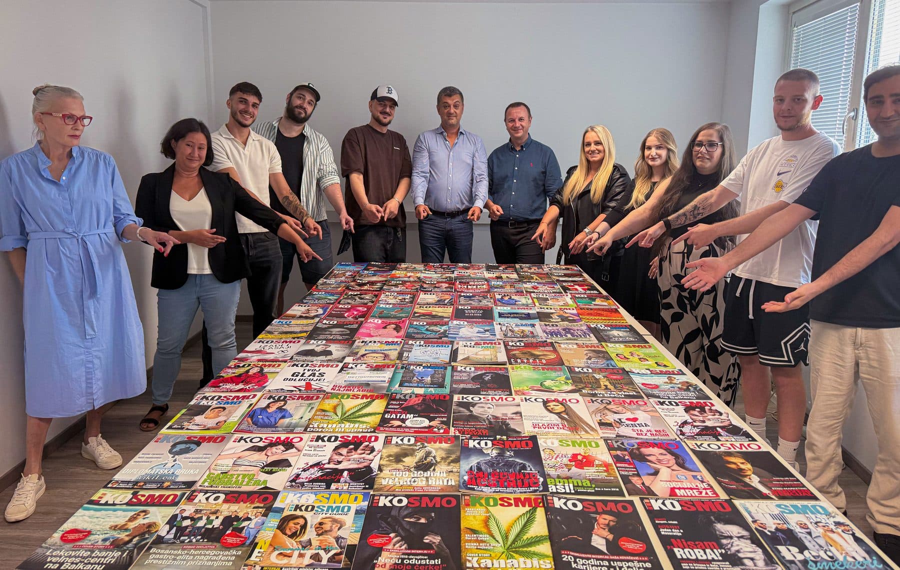 Das KOSMO-Team steht um einen Tisch, auf dem Dutzende KOSMO-Titelseiten ausgelegt sind, und zeigt auf sie
