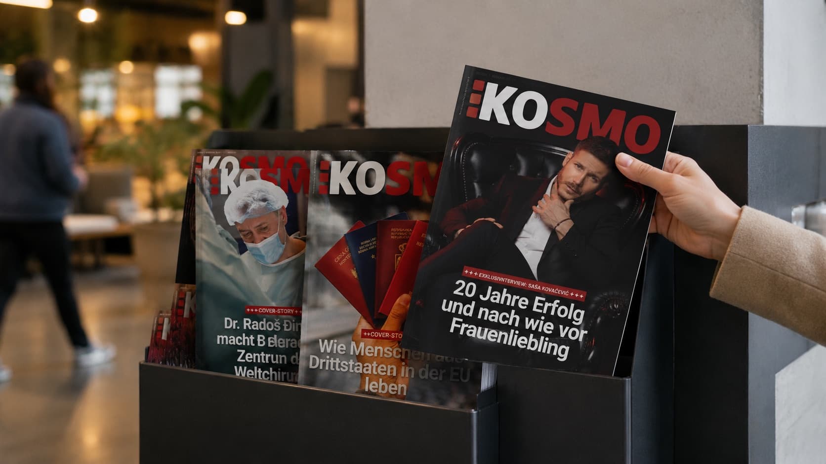 Eine Hand nimmt ein KOSMO-Heft aus einem Selbstbedienungsaufsteller